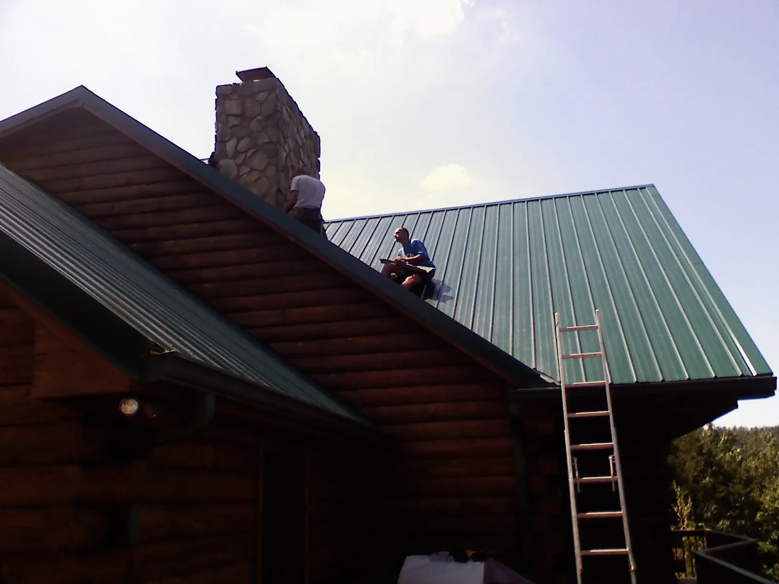 Roofers installing green metal panels beside a stone chimney on a log home in Hill City